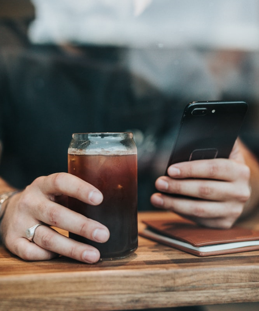 Man in a coffee shop holding mobile phone with one hand and a coffee with the other.